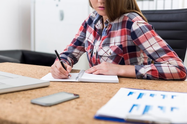 Vue rapprochée d'une jeune femme en chemise à carreaux qui écrit une note dans son cahier tout en étant assise à une table en bois avec un ordinateur portable et un téléphone dessus.
